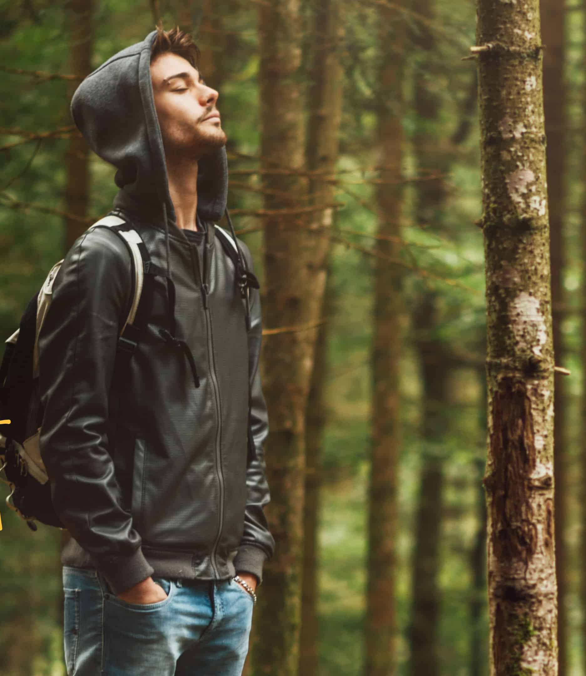 Junger Mann mit Kapuze und Rucksack steht mit geschlossenen Augen im Wald